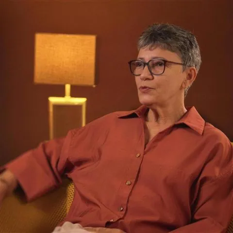 Une invitée en chemise rouille répond à une interview, assise dans un fauteuil devant une lampe allumée