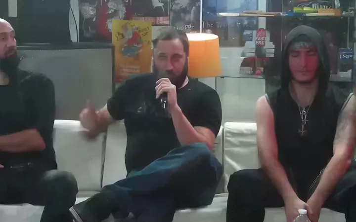 Deux hommes assis sur un canapé blanc lors d'une interview, l'un tient un micro