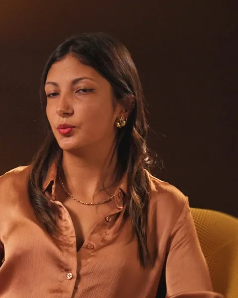 Une femme en chemisier satiné répond à une interview, assise dans un fauteuil jaune près d'une lampe chaude