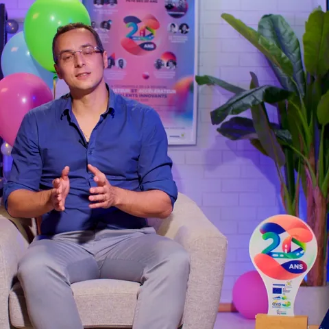 Un intervenant en chemise bleue assis dans un fauteuil, dans un décor de fête avec ballons et trophée 20 ans